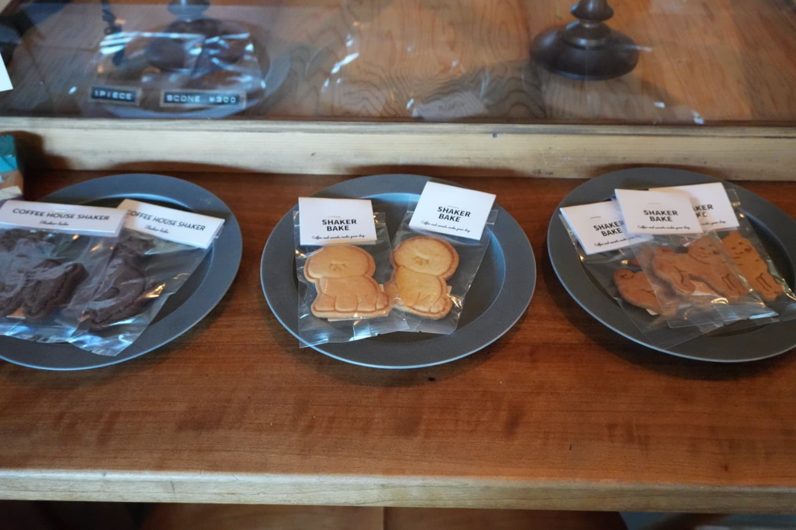 カウンターに並ぶ店のロゴ入りの焼き菓子。動物のかたちの「SHAKER BAKE」と「COFFEE HOUSE SHAKER」の袋が皿に載り、奥のショーケースには「SCONE ¥300」の表示(2026年9月22日訪問)