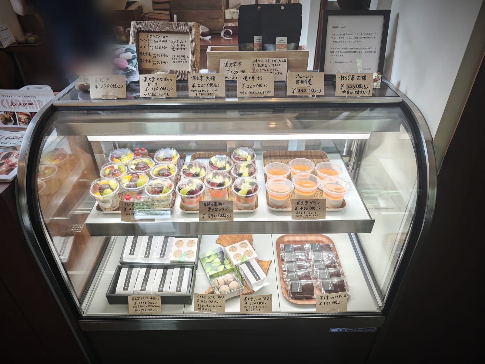 ショーケース全体。上段は季節の生菓子とプリン、下段は黒文字ようかんやくろもじしぐれなど手みやげ向きの品が並びます。奥のスタッフの方はぼかしています(2026年9月12日・編集部撮影)