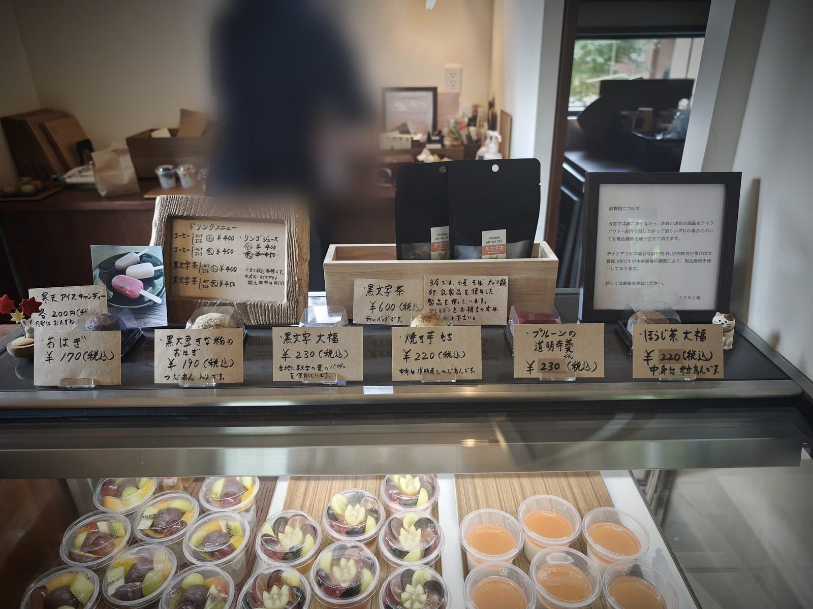 ショーケース上の価格札。おはぎ170円、黒文字大福230円、ほうじ茶大福220円など、この日の税込価格が並んでいました。奥のスタッフの方はぼかしています(2026年9月12日・編集部撮影)